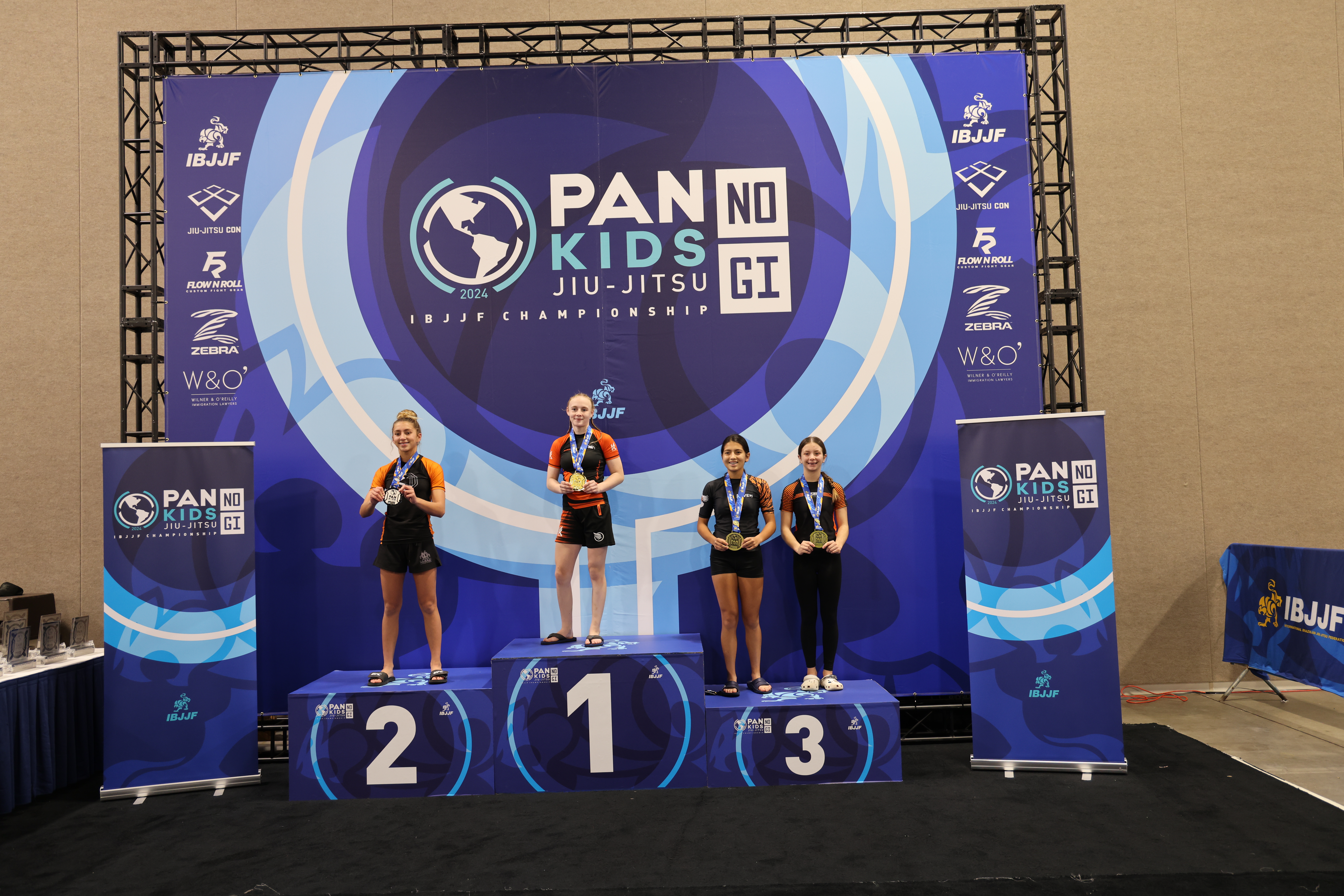 Mynd af verðlaunapalli frá Pan Kids IBJJF No-Gi Championship 2024. Efsta þrep pallsins er skipað meistara flokksins. Merkingar mótsins sjást í bakgrunni: PAN KIDS NO-GI IBJJF CHAMPIONSHIP.