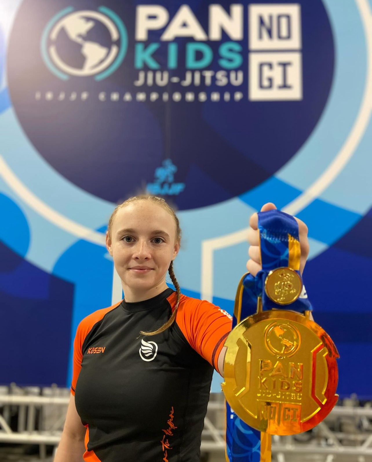 Mynd af gullverðlaunum tekin á IBJJF Pan Kids No-Gi Championship 2024. Á verðlaunapeningnum er auðkenni Pan Kids IBJJF Championship. Bakgrunnstjald mótsins, PAN KIDS NO-GI IBJJF CHAMPIONSHIP, sést í bakgrunni.