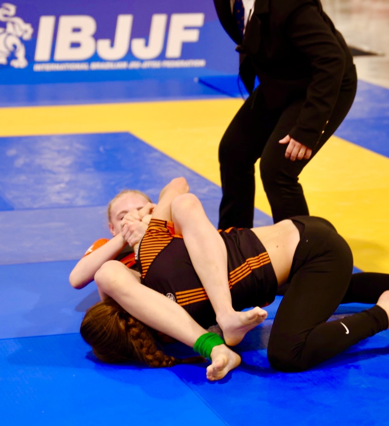 Frekari keppnismynd úr keppnistöflu IBJJF Pan Kids No-Gi Championship 2024. Viðfangið er sýnt í yfirfærslu úr yfirráðum í uppgjafartak. Merkingar IBJJF sjást á bakgrunnstjaldi keppnismottunnar.